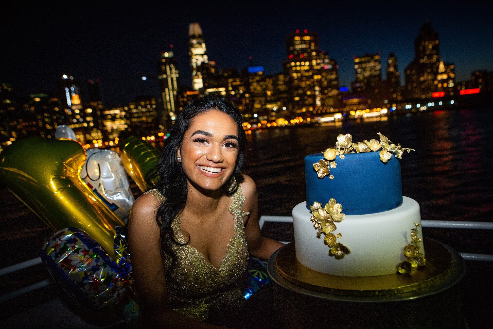 Smiling woman in a gold dress celebrating a milestone birthday with a decorated cake and balloons on a boat against a city skyline at night.